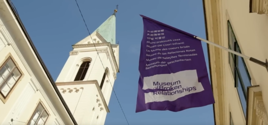 A clear blue sky with a church tower in the background. In front, a purple flag with white text hangs from a building. The flag says ‘Museum of Broken Relationships,’